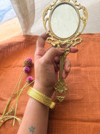 A wide gold cuff bracelet with a multi-layered ribbed texture worn on a wrist holding an ornate vintage hand mirror over a burnt orange fabric.
