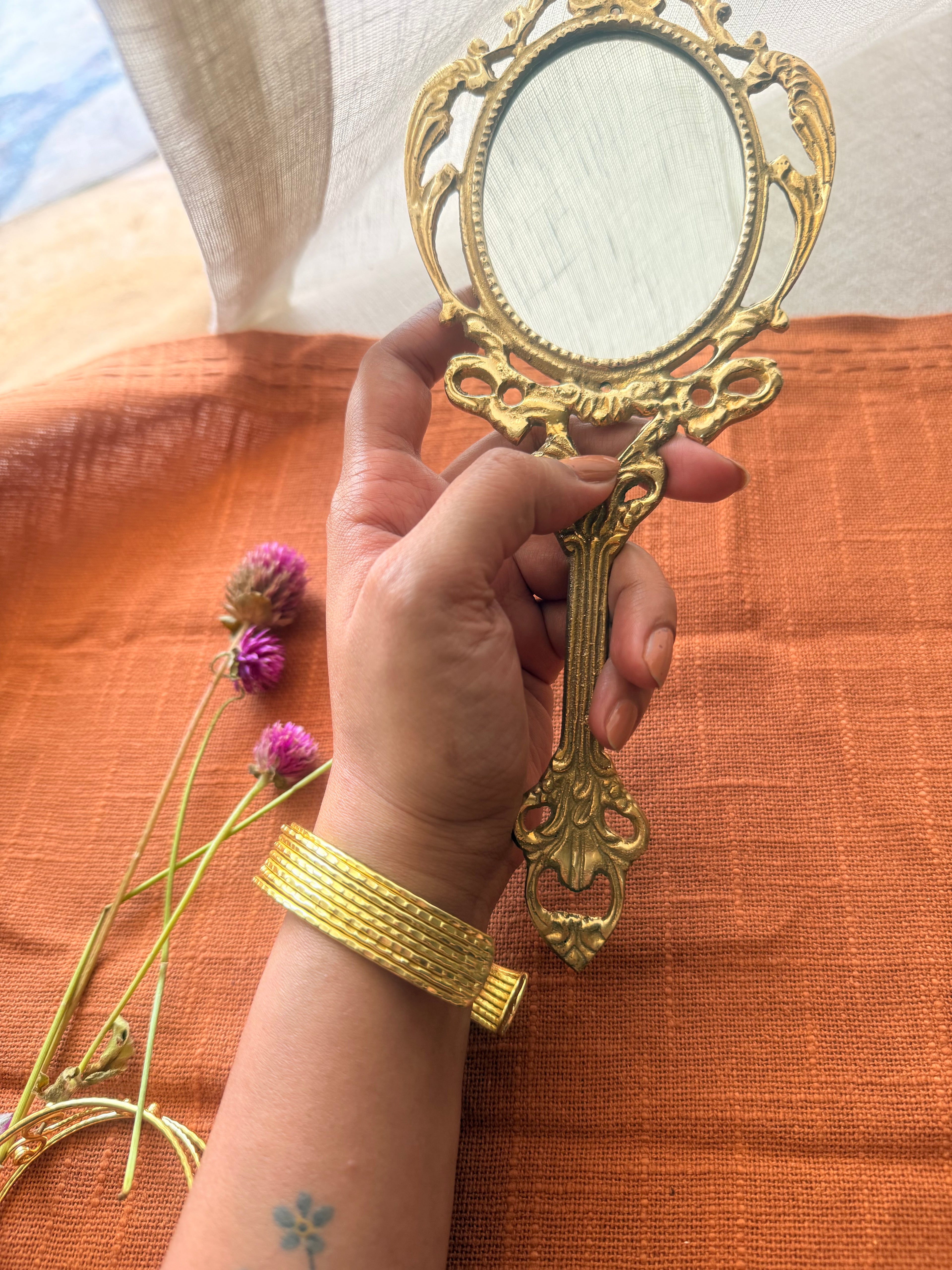 A wide gold cuff bracelet with a multi-layered ribbed texture worn on a wrist holding an ornate vintage hand mirror over a burnt orange fabric.