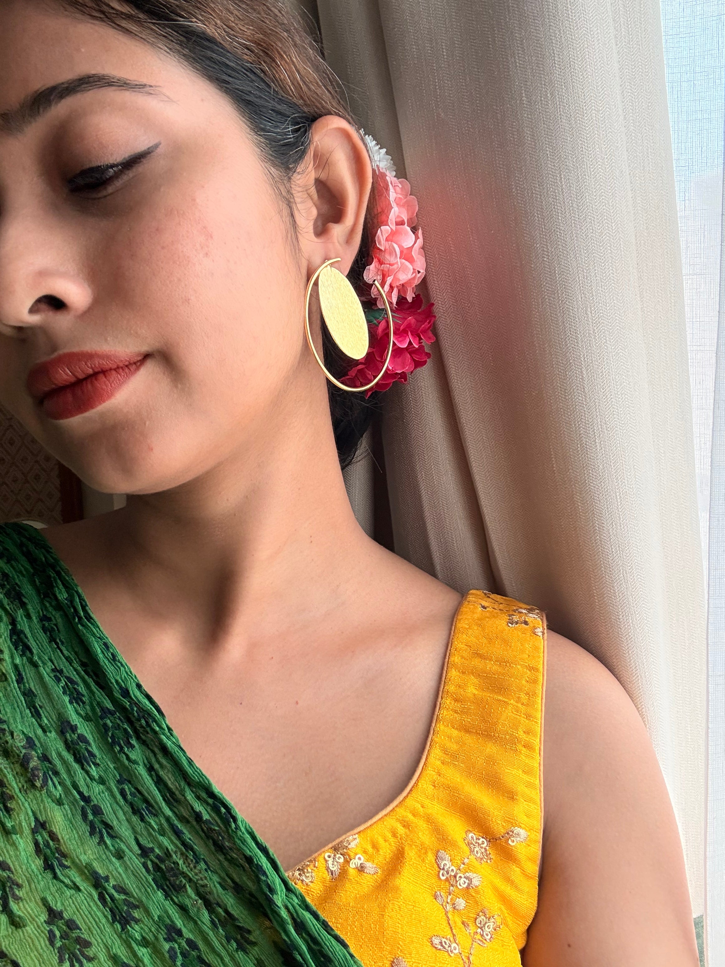 Close-up of woman wearing Sama Hoops with oval disc, dressed in a green saree and mustard blouse, looking downwards, pink flowers in hair