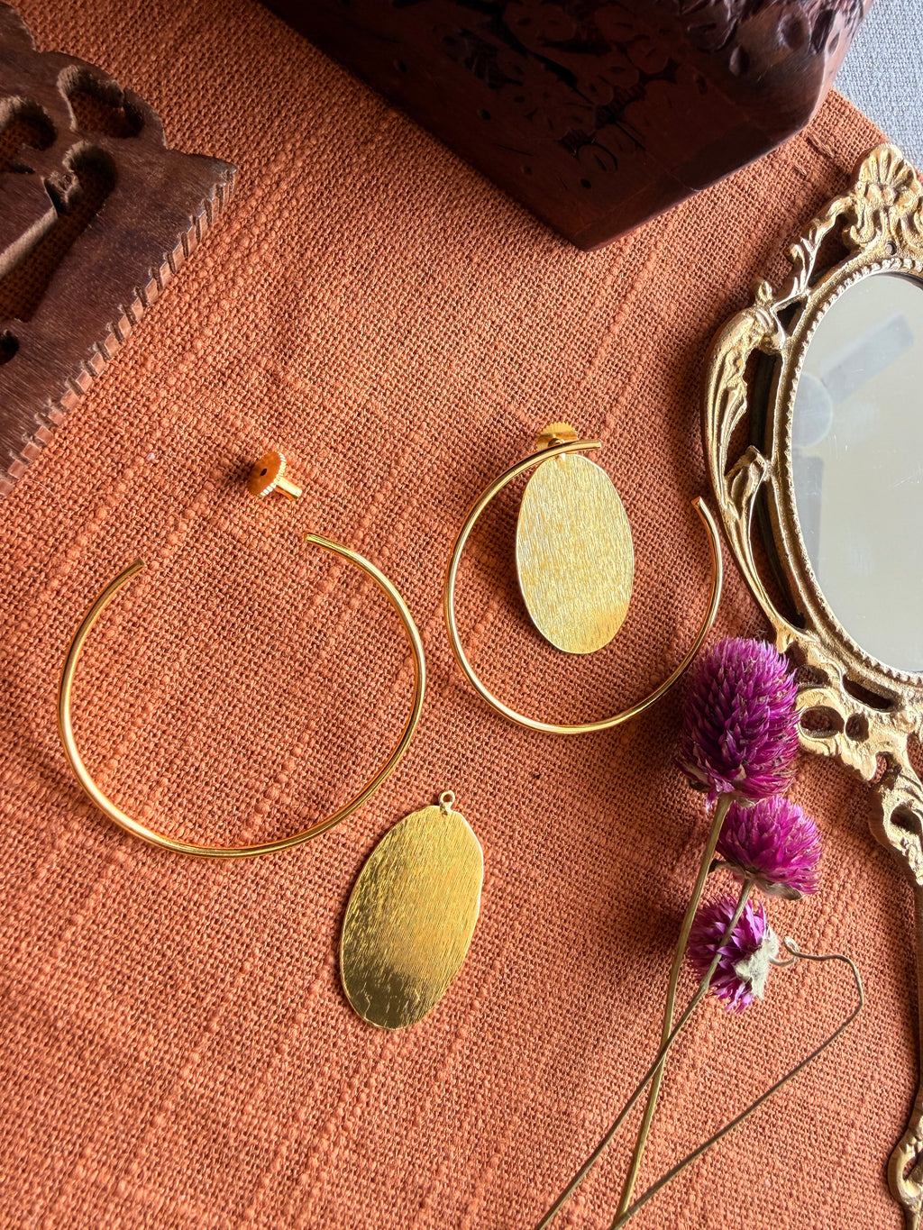 Sama Hoops flatlay showing hoop and detached oval disc separately on terracotta linen, with antique mirror and dried purple flowers