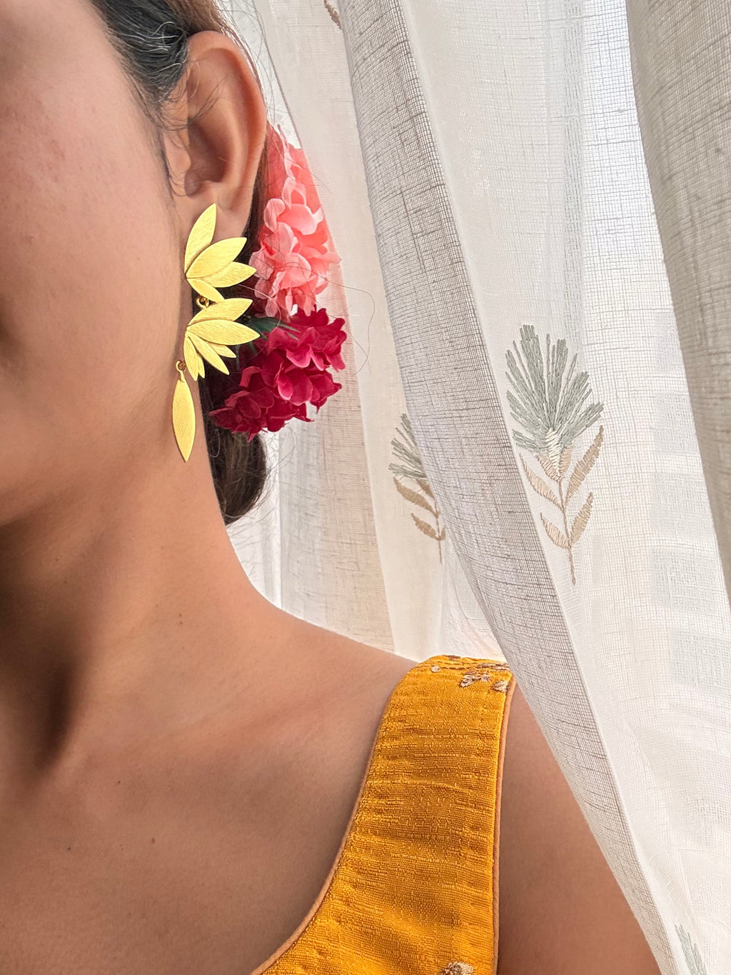 view of woman wearing Apsara danglers with pink and red flowers in hair, mustard embroidered blouse, sheer floral curtain in background
