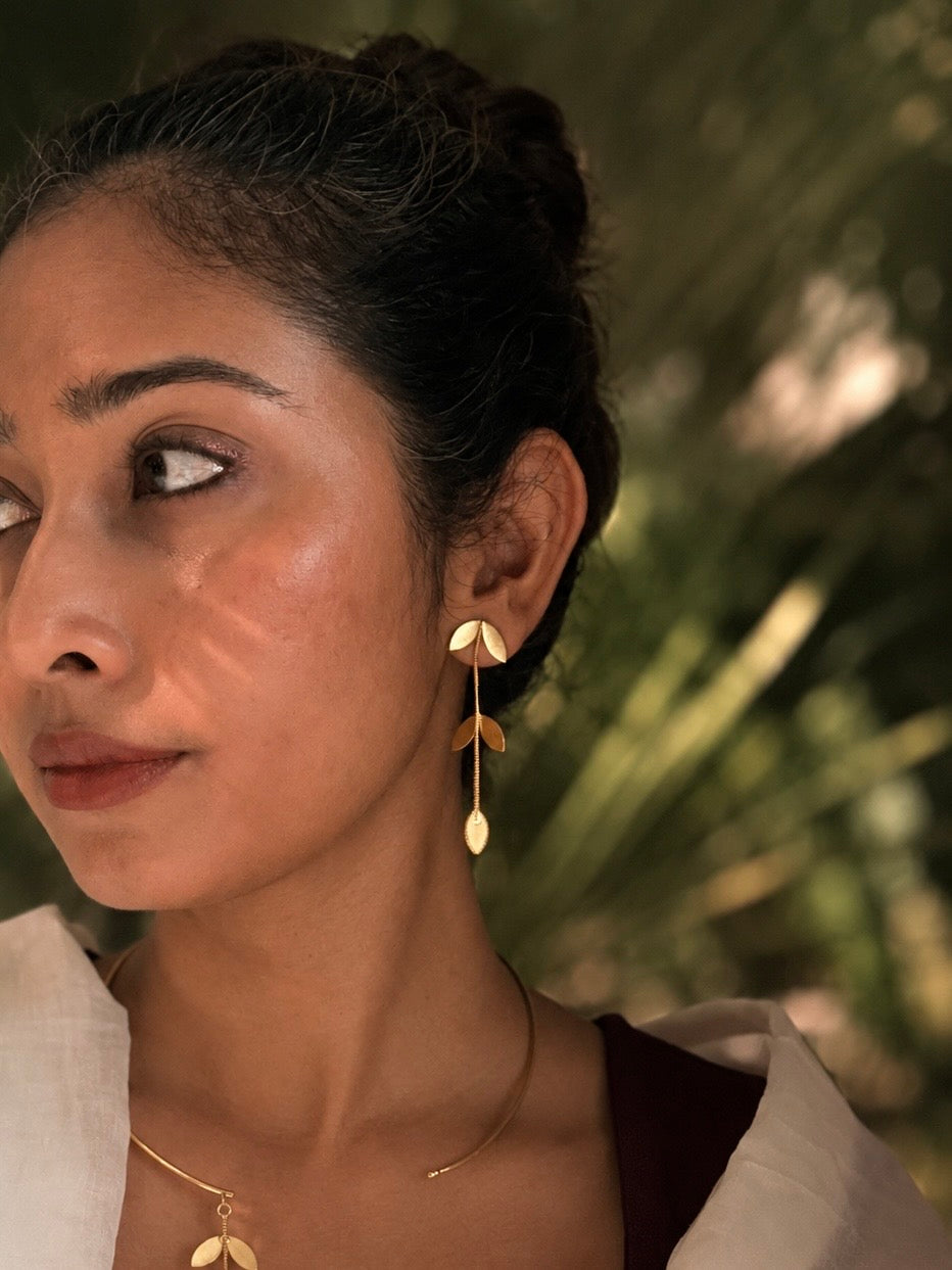 Close-up of woman wearing Pallavi earrings with hair in an updo, gold leaf cascade visible against her neck alongside the Pallavi choker necklace, dark botanical background, warm natural light