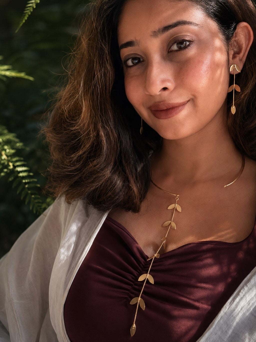 Woman wearing a maroon top with a gold necklace and earrings against a green leafy background