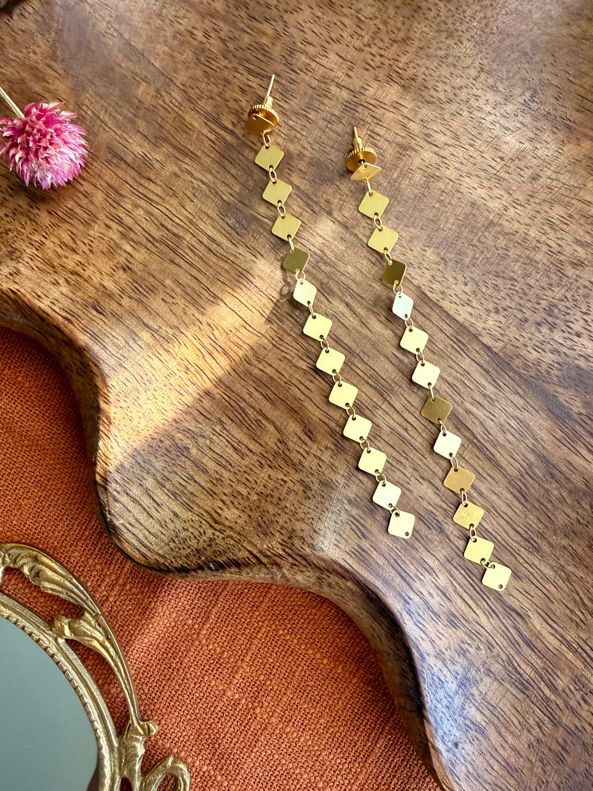 Pair of long gold Aabha Danglers with small diamond-shaped links, laid on a carved wooden tray beside a dried pink flower and an antique brass mirror