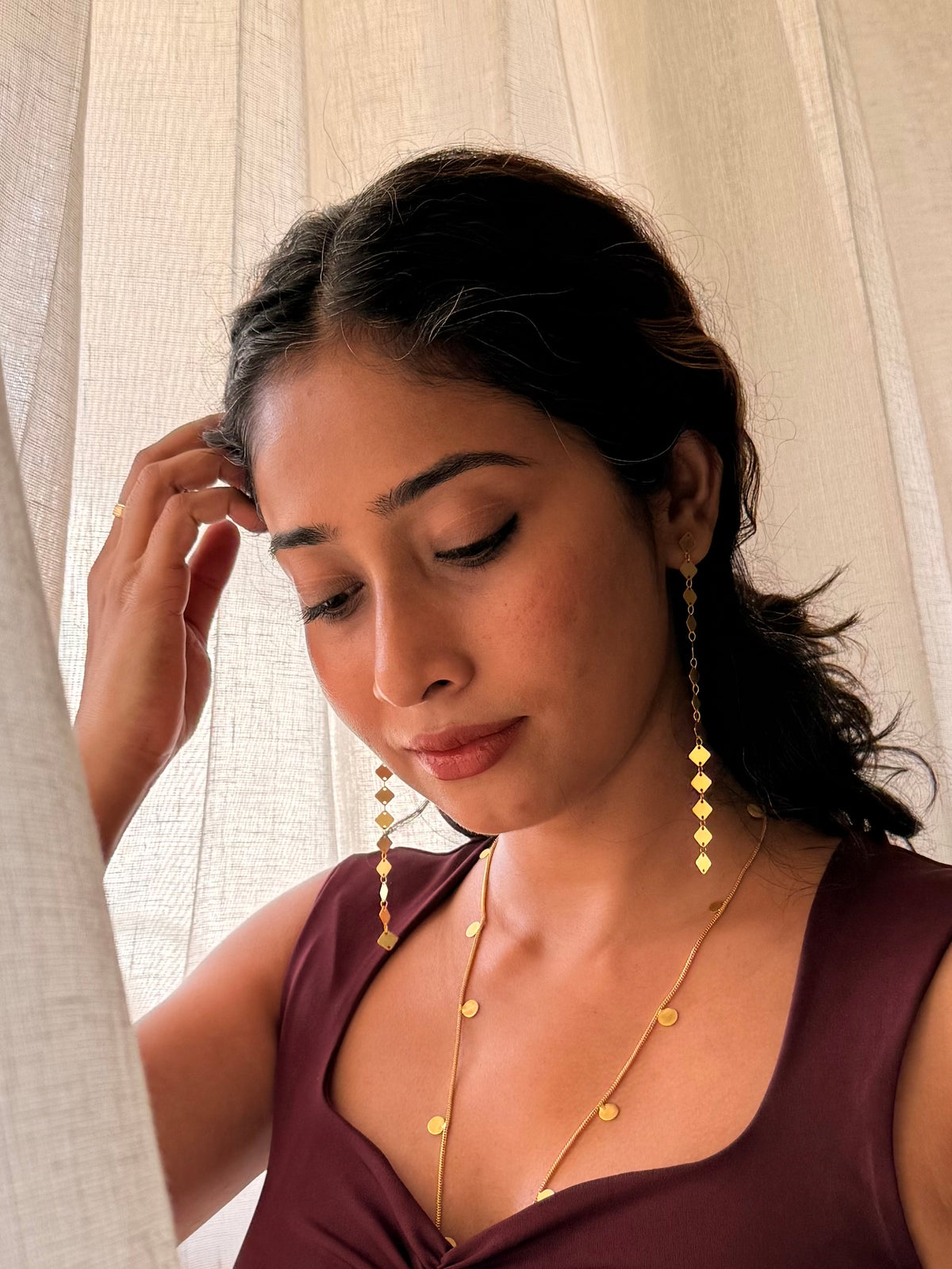 Woman looking down with one hand in her hair, wearing Aabha Danglers and a fine gold necklace in front of sheer white curtains