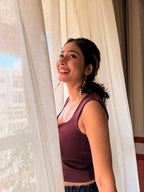 Smiling woman in a maroon top standing by sheer curtains in soft sunlight, wearing long gold Aabha Danglers