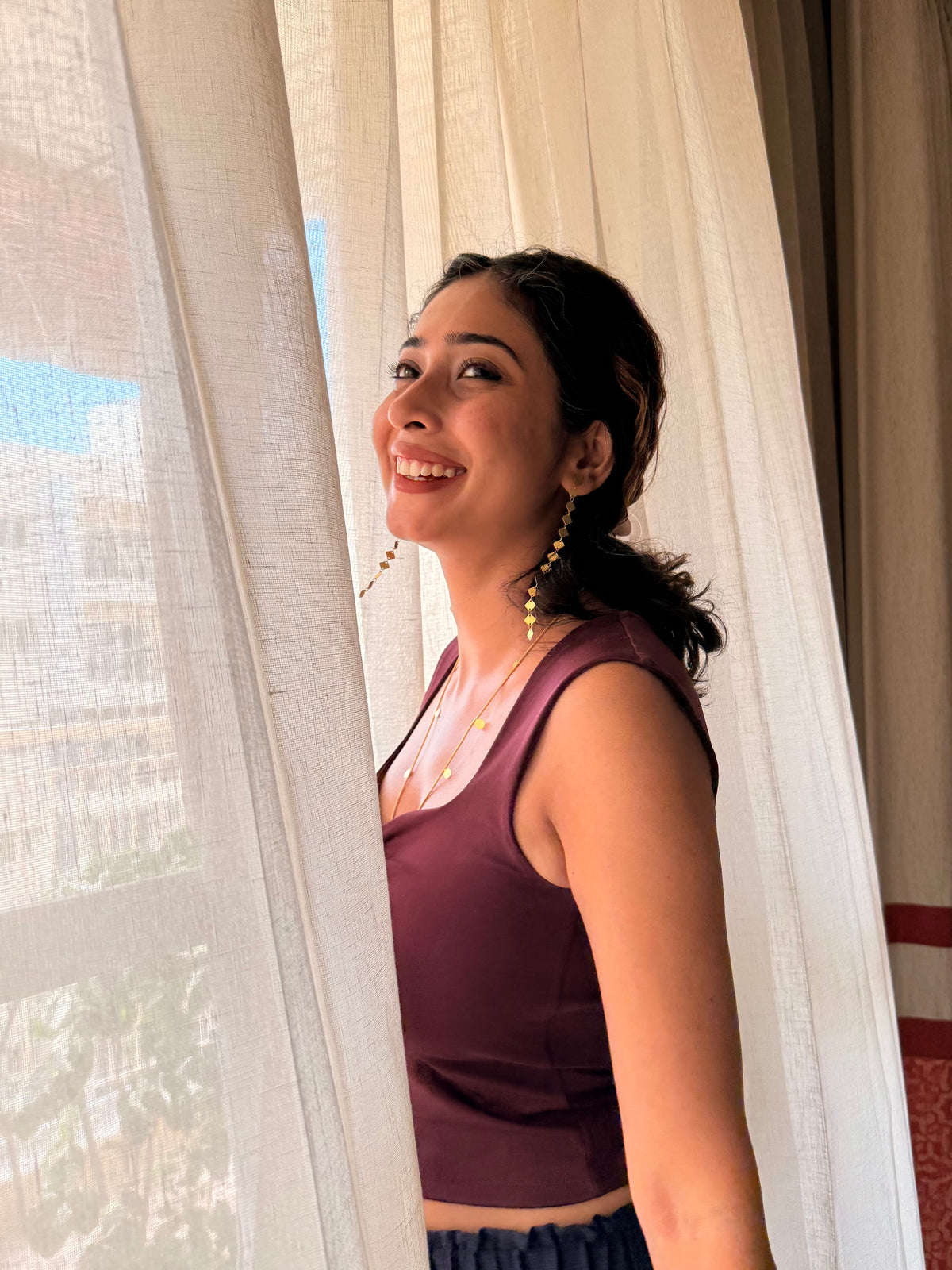 Smiling woman in a maroon top standing by sheer curtains in soft sunlight, wearing long gold Aabha Danglers
