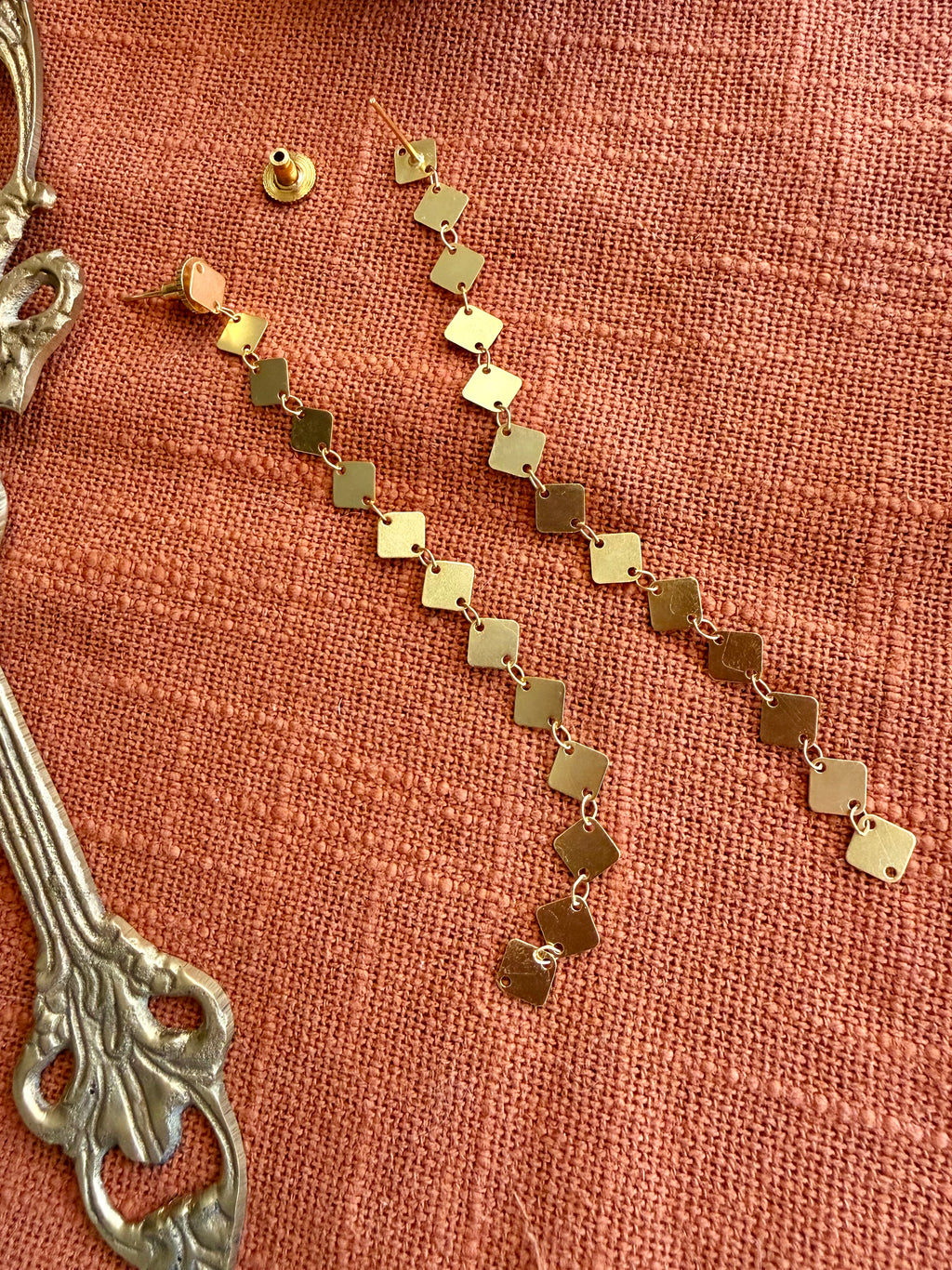 Close-up of Aabha Danglers laid flat on rust-orange linen beside an ornate brass handle, showing the diamond-shaped links and earring back