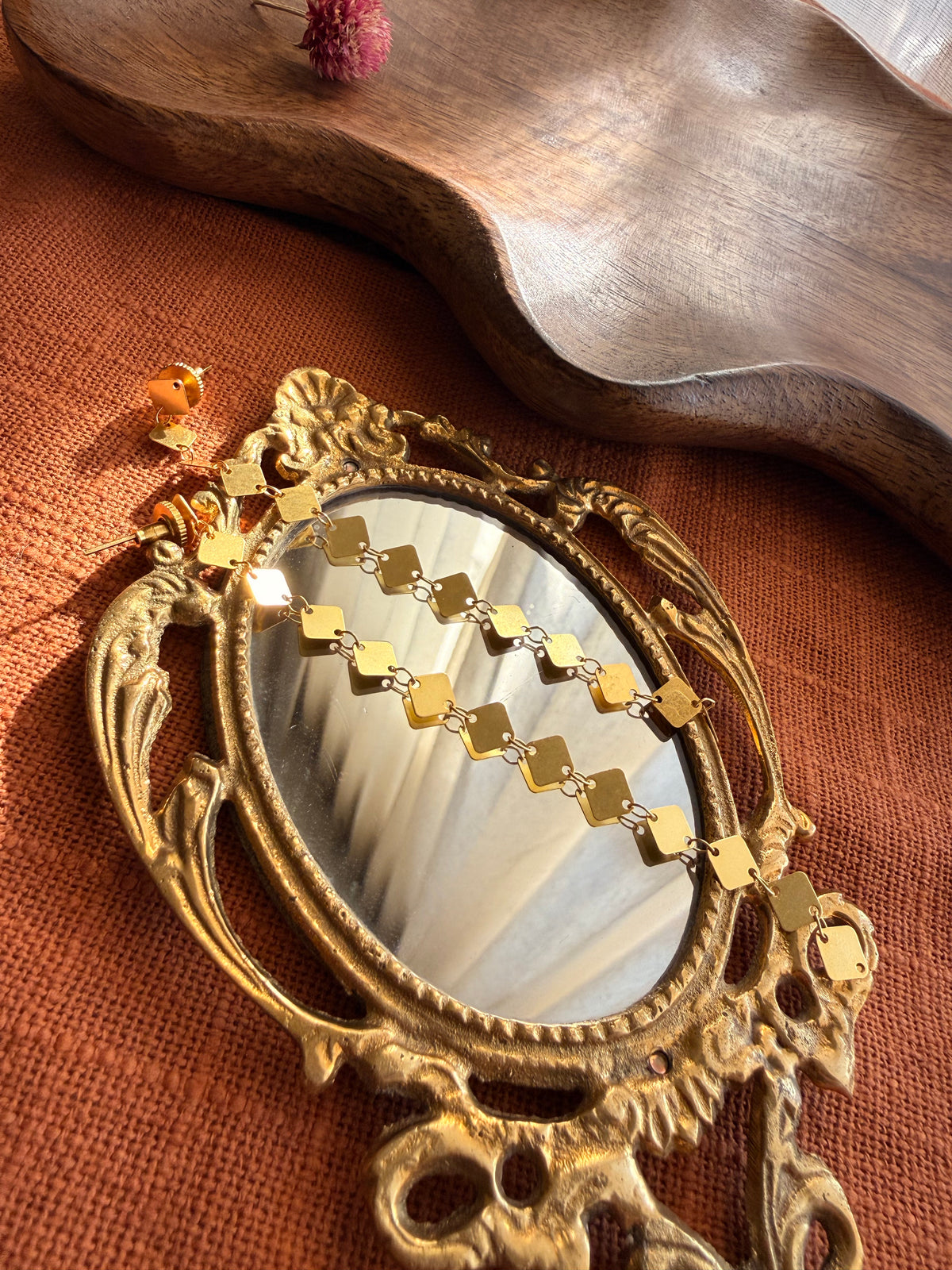 Aabha Danglers resting across an antique brass hand mirror on rust linen, with a wooden tray and dried flower behind
