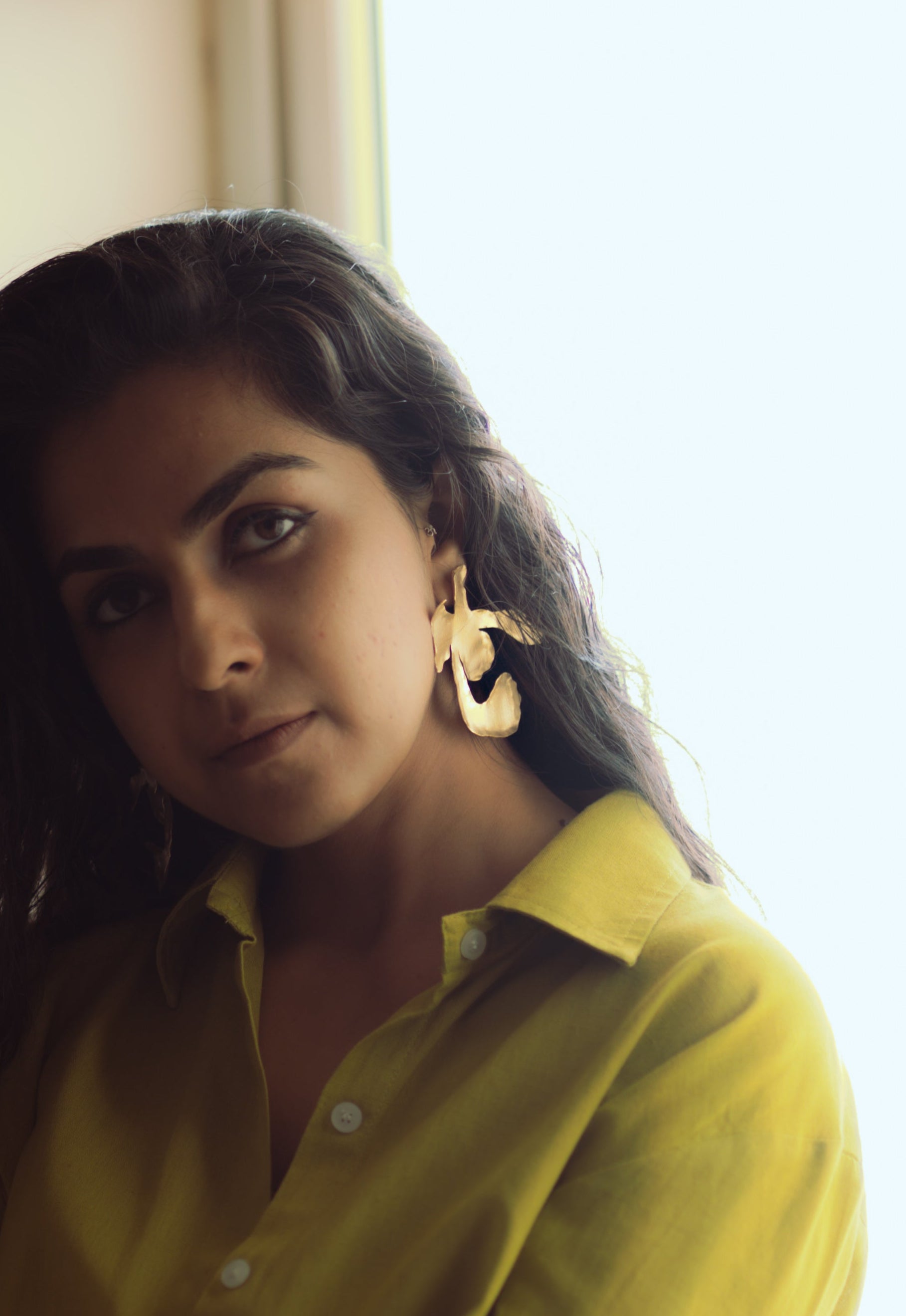 Closeup of Woman in a light green shirt sitting by a window, wearing Ankur Twist Earrings.