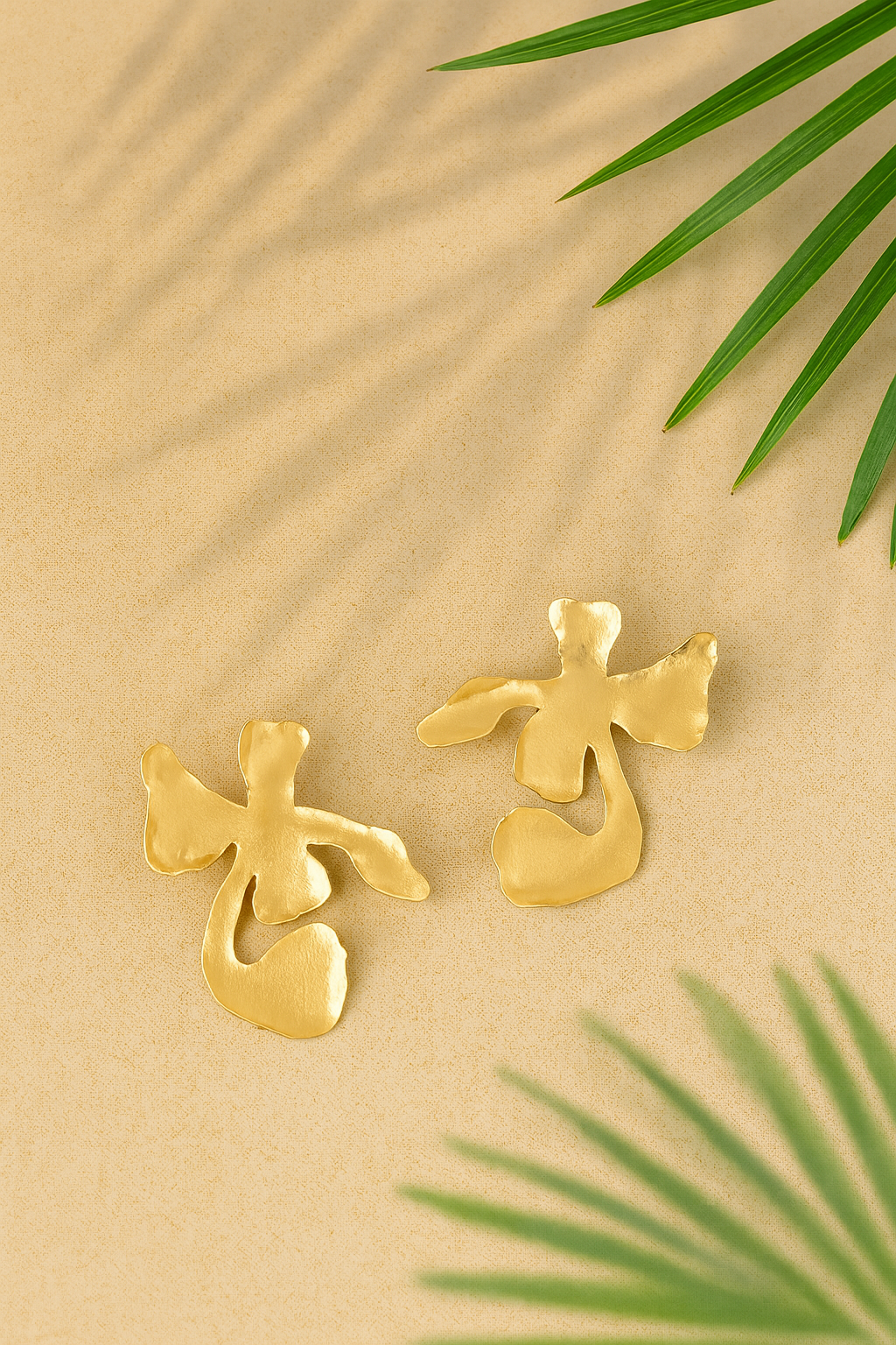 Gold plated floral shaped Ankur Twist Earrings on a beige background with green leaves