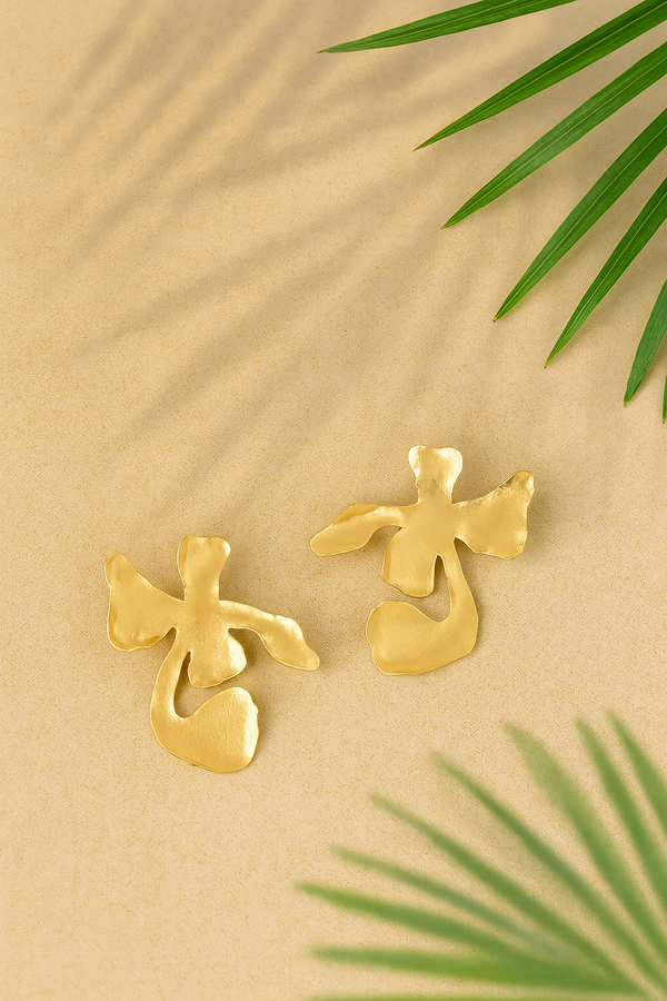 Gold plated floral shaped Ankur Twist Earrings on a beige background with green leaves