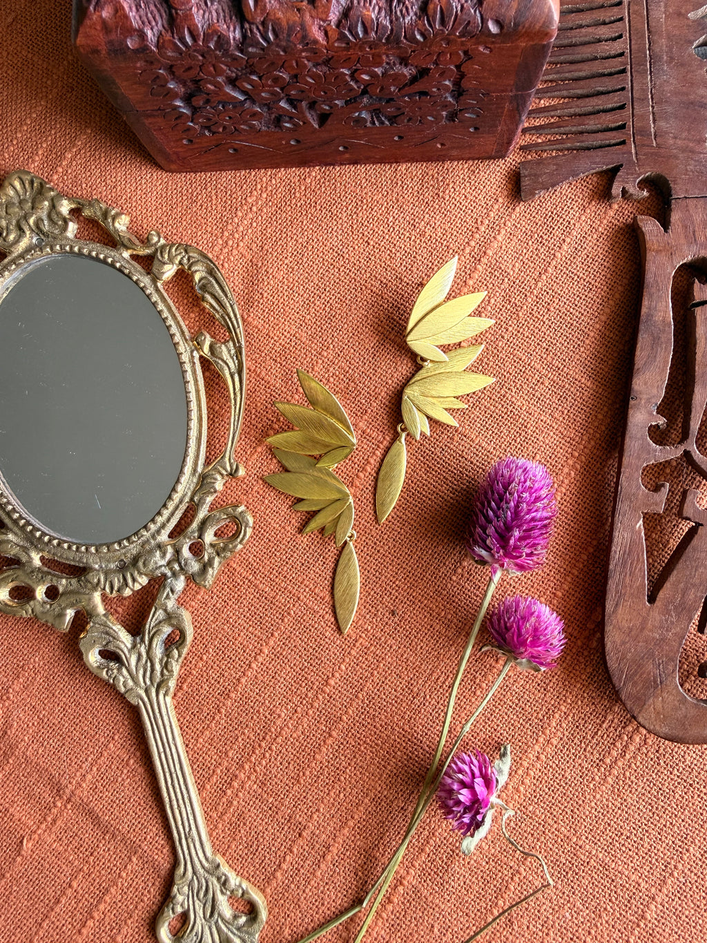 Gold-plated brass Apsara danglers flatlay on terracotta fabric, showing petal cluster and feather drop separated, styled with vintage mirror and dried purple Globe Amaranth flowers