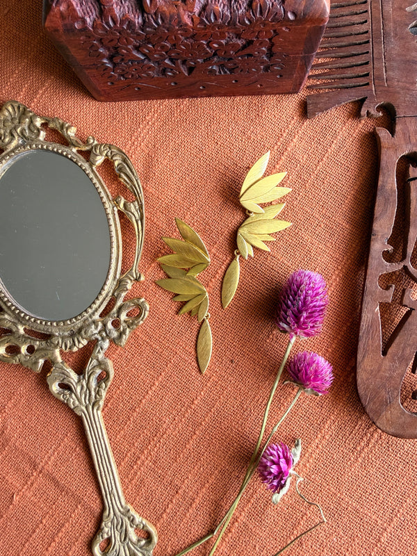 Gold-plated brass Apsara danglers flatlay on terracotta fabric, showing petal cluster and feather drop separated, styled with vintage mirror and dried purple Globe Amaranth flowers
