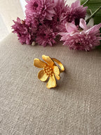 Ban Gulab gold flower-shaped ring with a textured center and polished band, displayed on a beige linen surface with pink flowers in the background.
