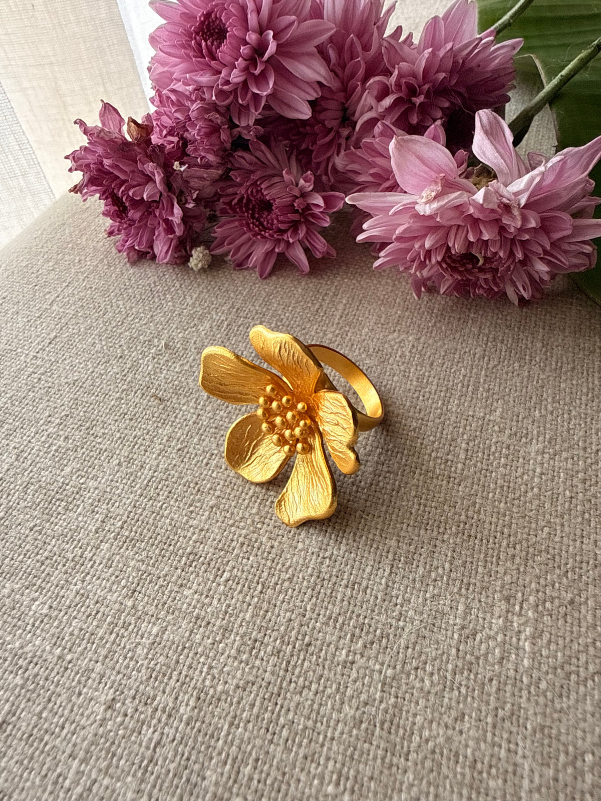 Ban Gulab gold flower-shaped ring with a textured center and polished band, displayed on a beige linen surface with pink flowers in the background.