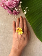 A woman's hand wearing ban gulab flower ring, styled with pink flowers and a green leaf on a beige linen background.