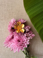 A cluster of pink chrysanthemum-like flowers with a ban gulab floral ring resting on top, arranged on a beige linen surface with a large green leaf in the upper right corner.