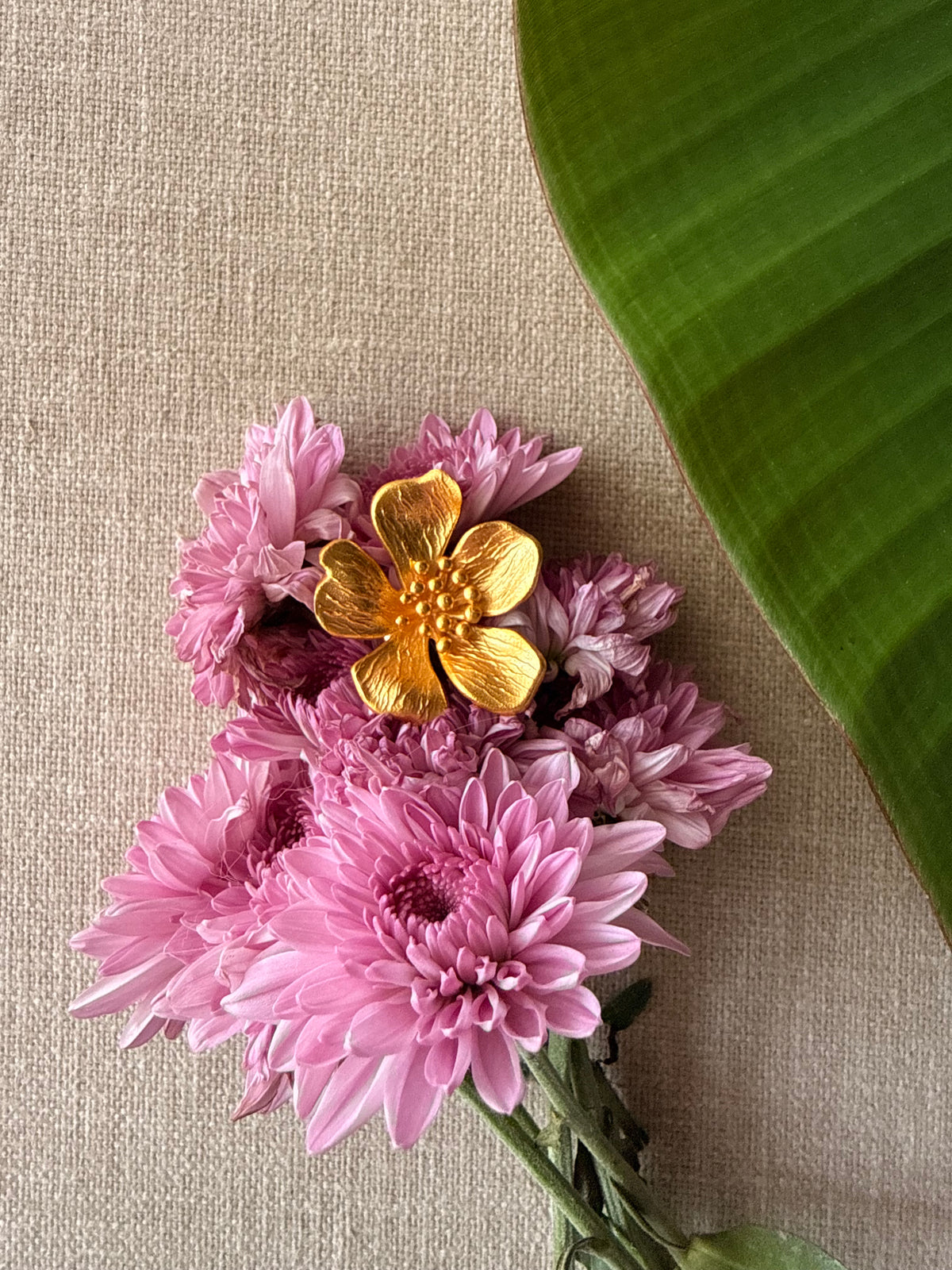 A cluster of pink chrysanthemum-like flowers with a ban gulab floral ring resting on top, arranged on a beige linen surface with a large green leaf in the upper right corner.