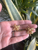 Gold bee-shaped earrings held in a hand with a plant in the background