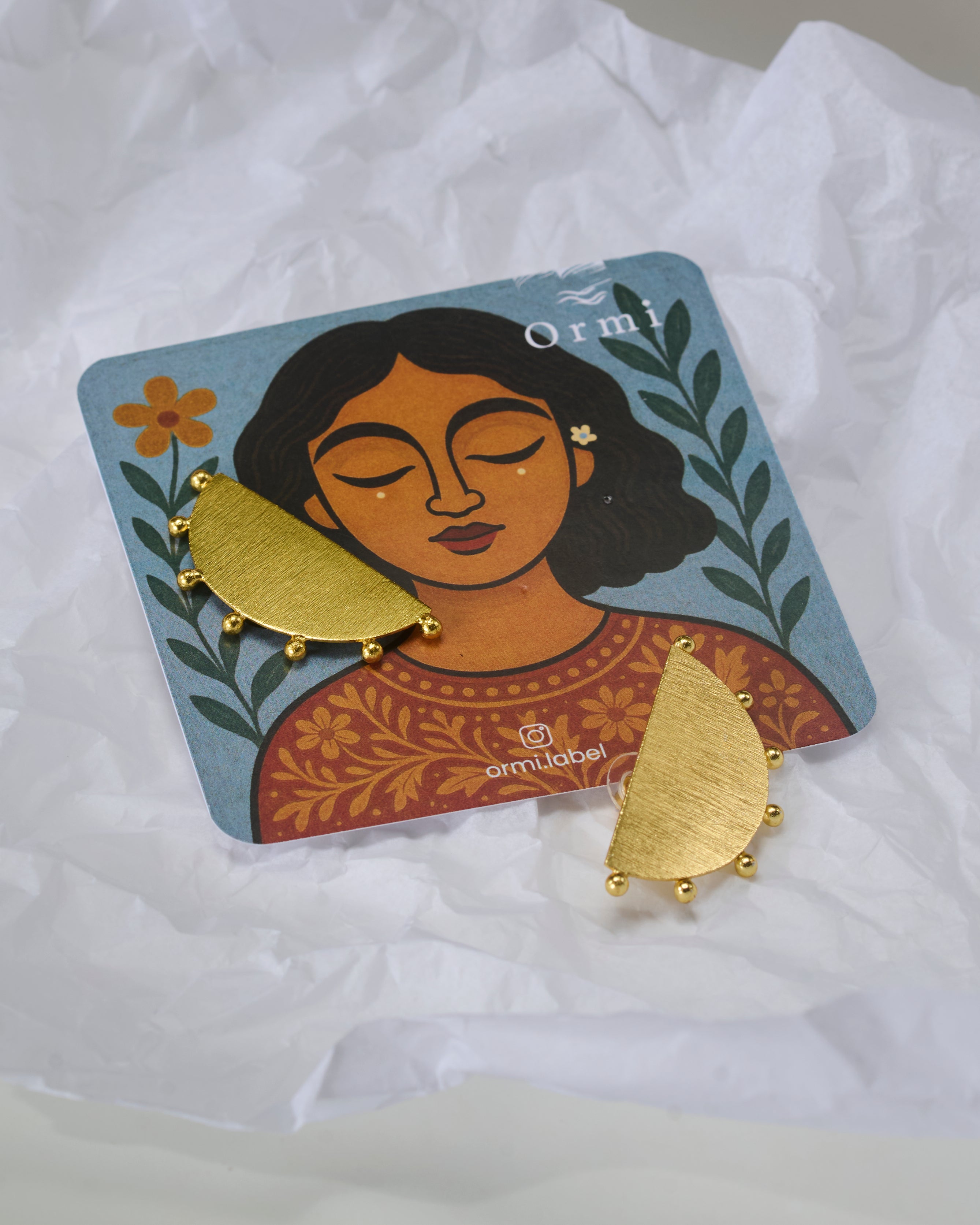 Gold toned half circular earrings with tiny circular details on a on a card with a woman's face, featuring the brand 'Ormi'.