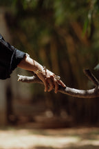 A hand resting on a tree branch, wearing Boond & Bel ring. Green blurry backgroud.