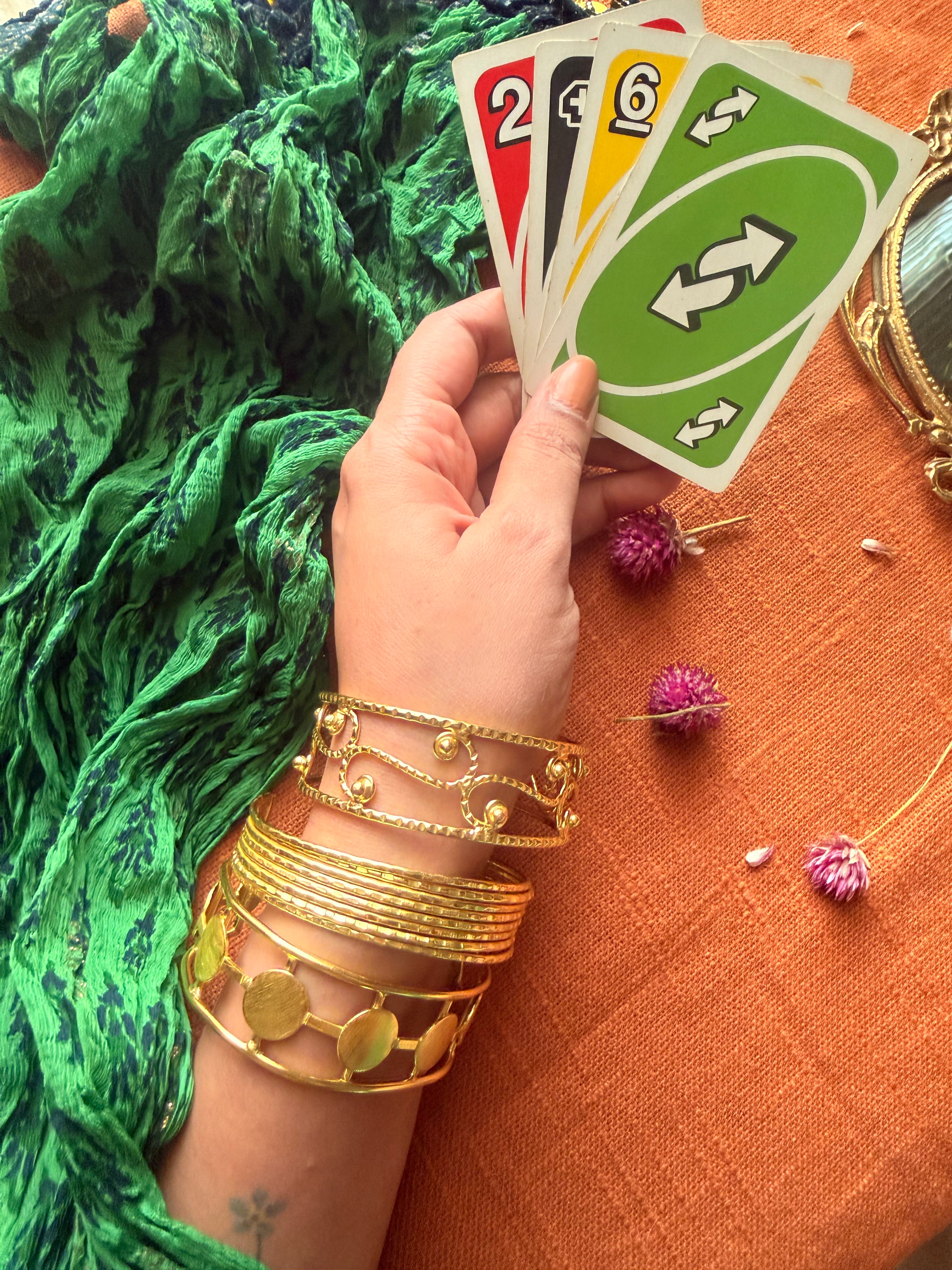 A lifestyle shot of a hand wearing a stack of three golden Ormi cuffs—the Filigree, Saj, and Chakra bracelets—while holding a hand of Uno cards. The background features a vibrant green dupatta and orange fabric with dried flowers, evoking a lived-in, everyday luxury vibe.