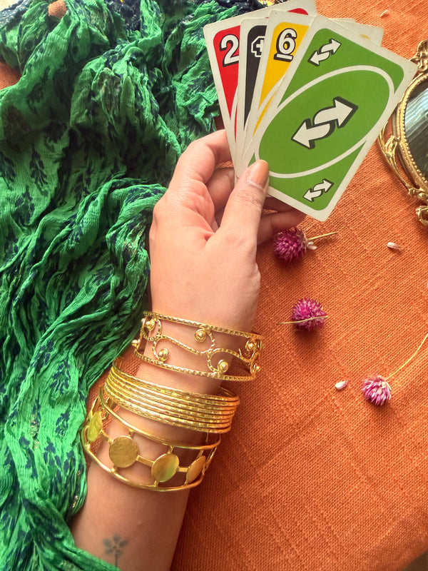 A lifestyle shot of a hand wearing a stack of three golden Ormi cuffs—the Filigree, Saj, and Chakra bracelets—while holding a hand of Uno cards. The background features a vibrant green dupatta and orange fabric with dried flowers, evoking a lived-in, everyday luxury vibe.
