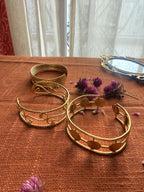 A warm, sunlit flatlay of the 'Golden Trio' bundle: the Saj, Filigree, and Chakra cuff bracelets arranged on an orange textured cloth next to dried purple flowers and a vintage-style gold mirror.