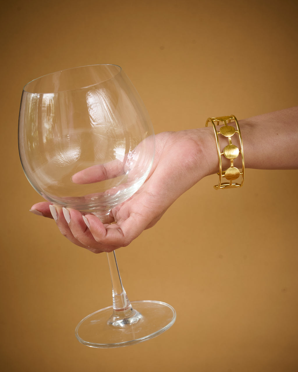 Hand holding a clear wine glass with a gold bracelet on a beige background
