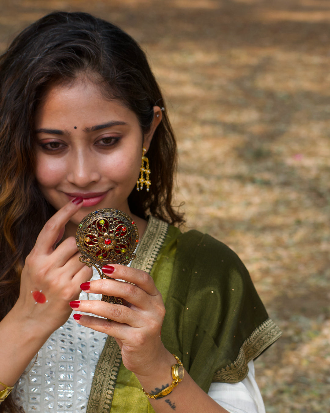 Woman wearing beautiful dangling earrings, holding a decorative mirror with a blurred natural background
