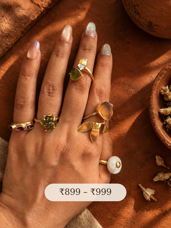 Hand wearing five handcrafted brass gemstone rings against terracotta, including Garnet Hydro, Green Moldavite, Peridot Hydro with crystal, Beer Quartz and Rainbow Moonstone, priced ₹899 to ₹999