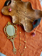 Deepmala Necklace laid flat as a long gold-toned chain with small scattered discs, on a wavy wooden tray beside a brass hand mirror and a dried pink globe amaranth, on rust linen