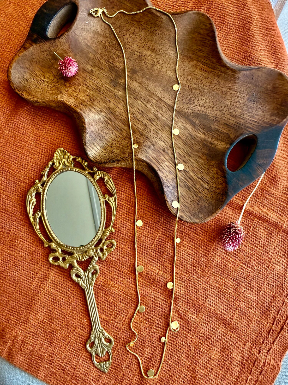 Deepmala Necklace laid flat as a long gold-toned chain with small scattered discs, on a wavy wooden tray beside a brass hand mirror and a dried pink globe amaranth, on rust linen