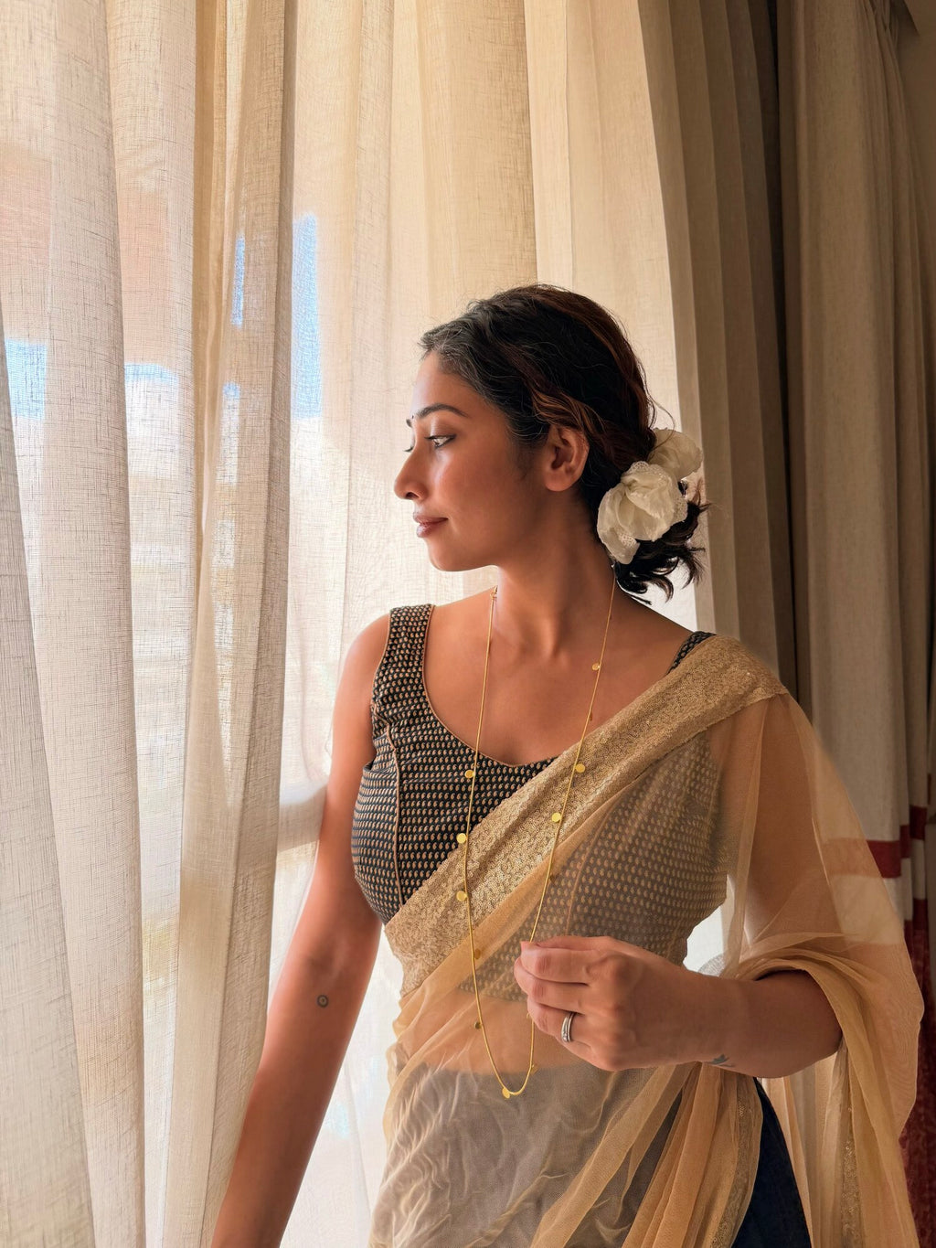 Woman in a sheer cream saree and grey blouse with a white flower in her hair, looking out a window with sheer curtains, wearing the Deepmala Necklace as a long single chain