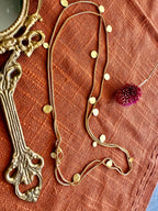 Close-up of the Deepmala Necklace doubled in loops on rust linen, small gold discs along the chain, beside a brass hand mirror and a dried pink flower