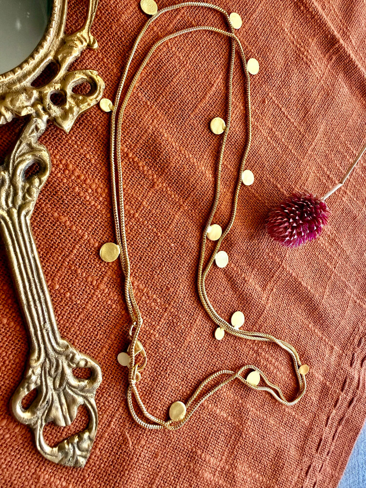 Close-up of the Deepmala Necklace doubled in loops on rust linen, small gold discs along the chain, beside a brass hand mirror and a dried pink flower