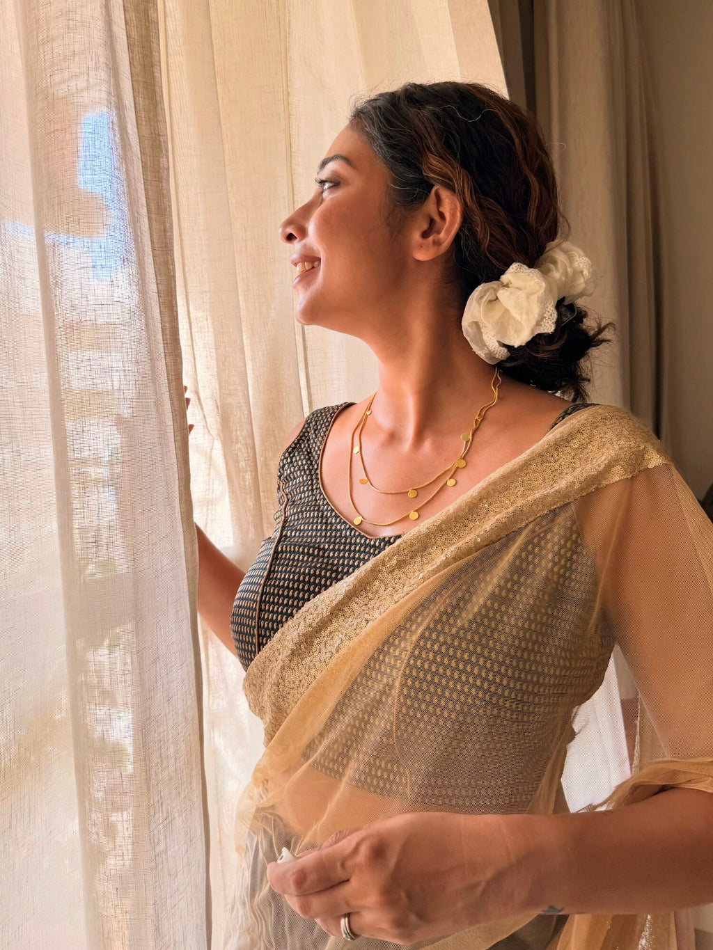 Side profile of a woman by a sheer curtain in a cream saree, smiling, wearing the Deepmala Necklace layered at the neck in multiple chain strands with small gold discs