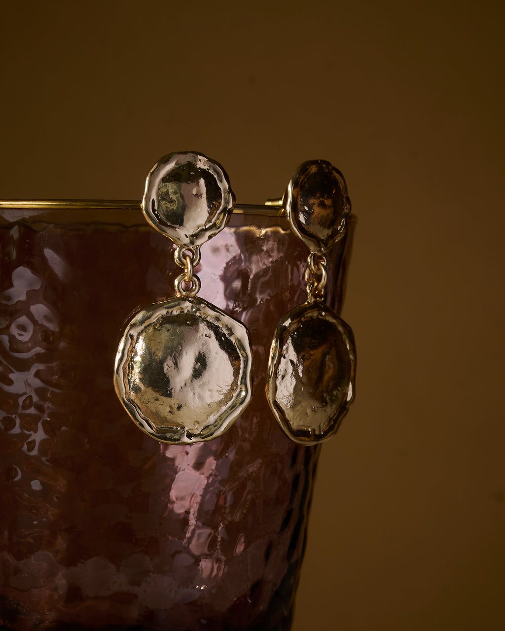 Close up of gold plated earrings with a textured organic finish, on a reflective surface.