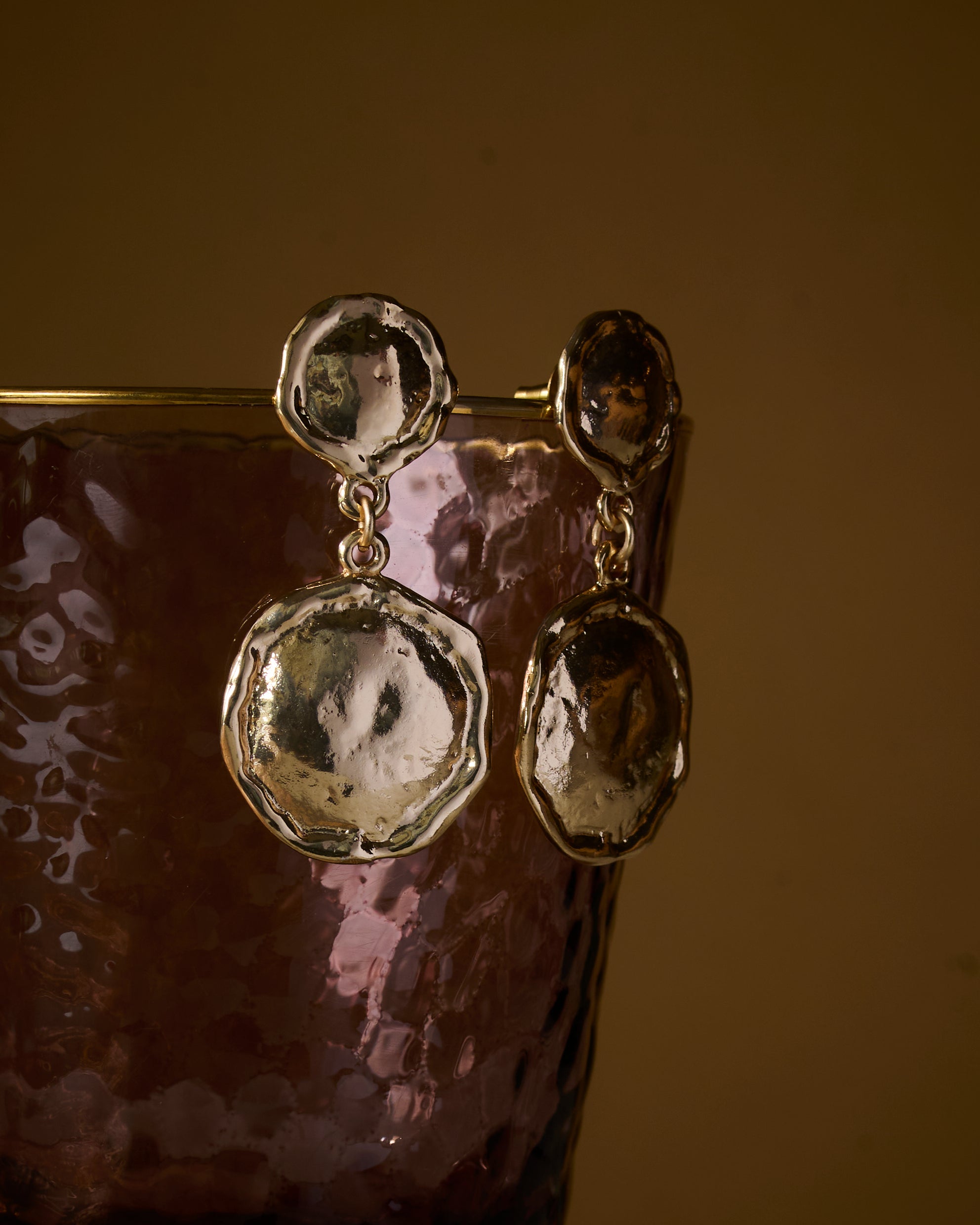 Close up of gold plated earrings with a textured organic finish, on a reflective surface.