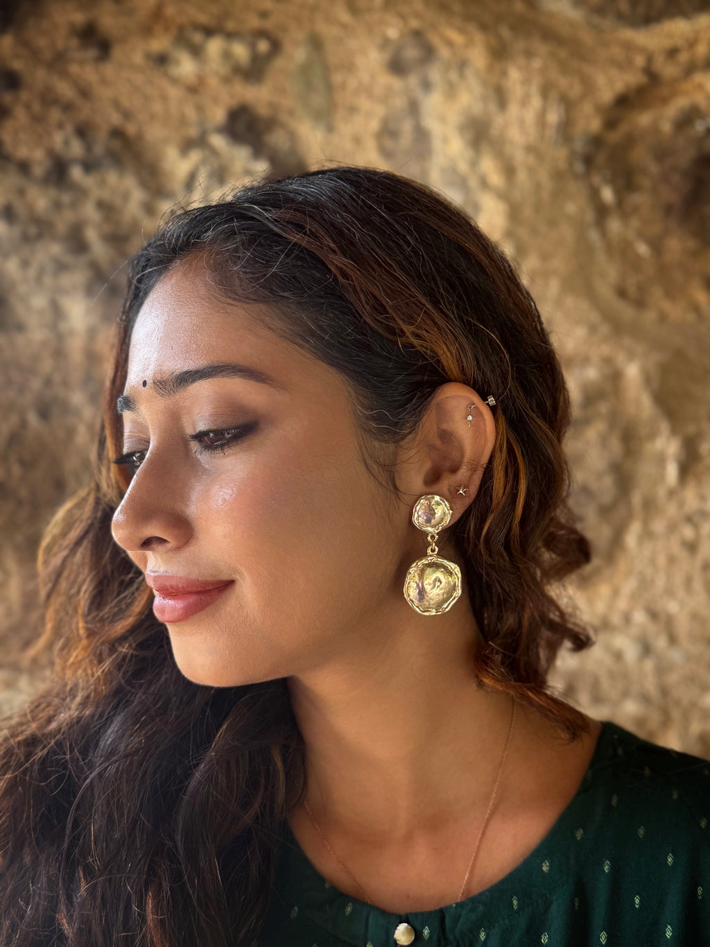 Woman wearing gold plated big and small circular disc shaped earrings against a textured wall background