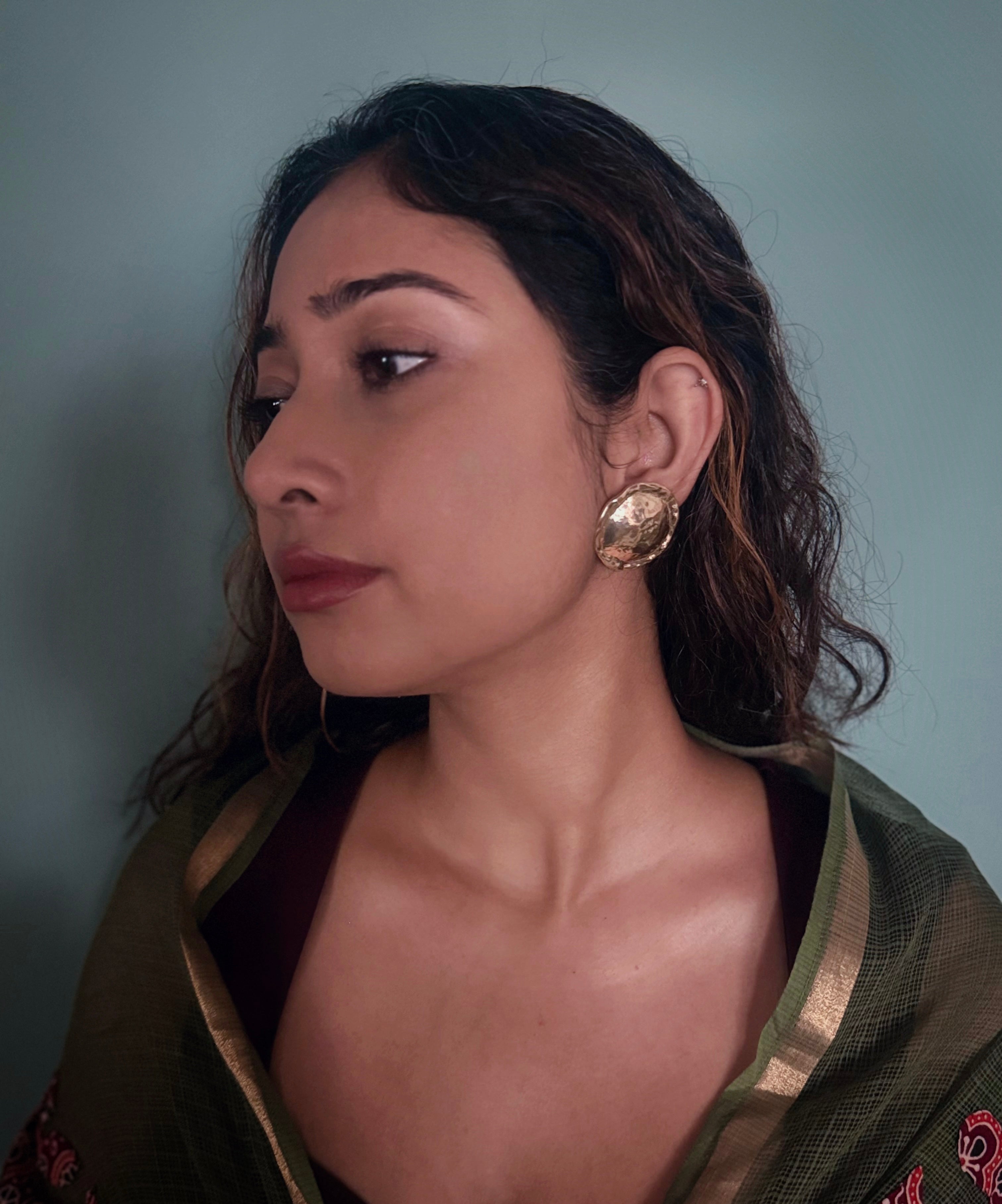 Woman with open messy hair, wearing gold plated circular textured earrings with a teal background