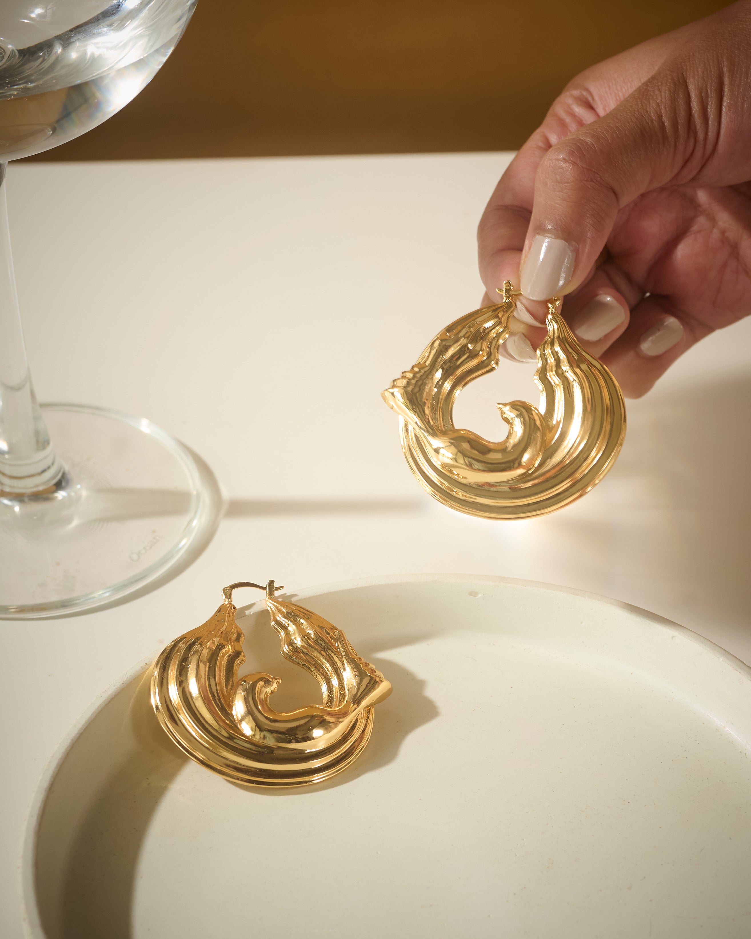 Gold earrings being held above a white surface with a glass of water in the background