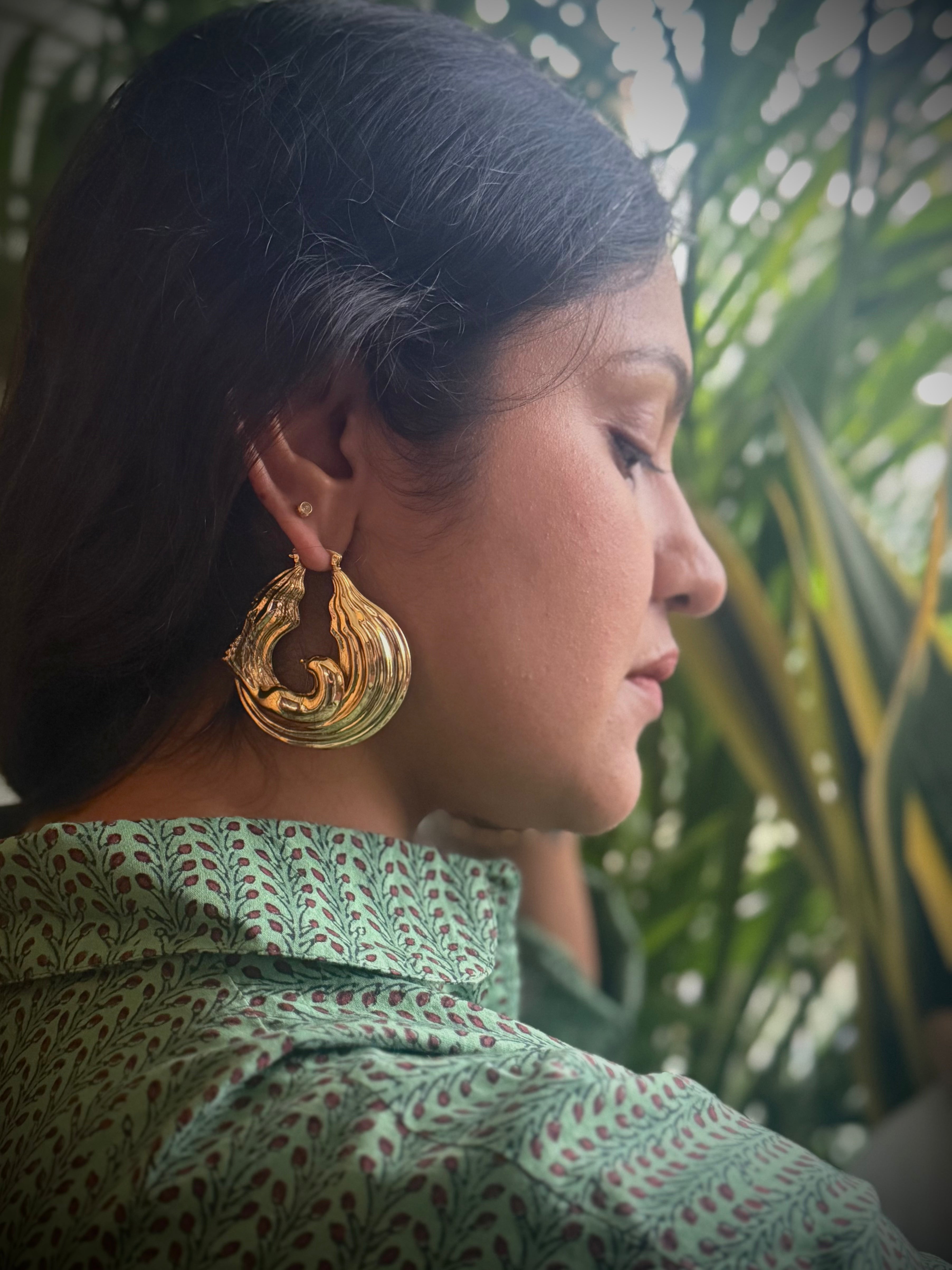 Woman wearing gold plated chunky hoop earrings with a green patterned top, surrounded by plants.
