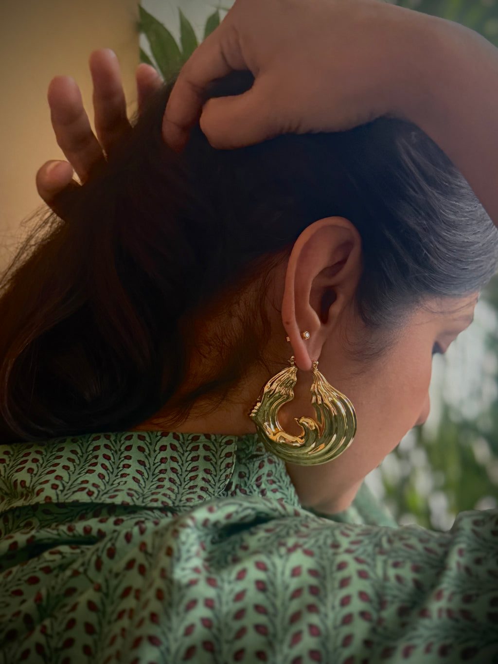 Woman holding back her hair, wearing gold plated chunky hoop earrings with a green patterned top, surrounded by plants.