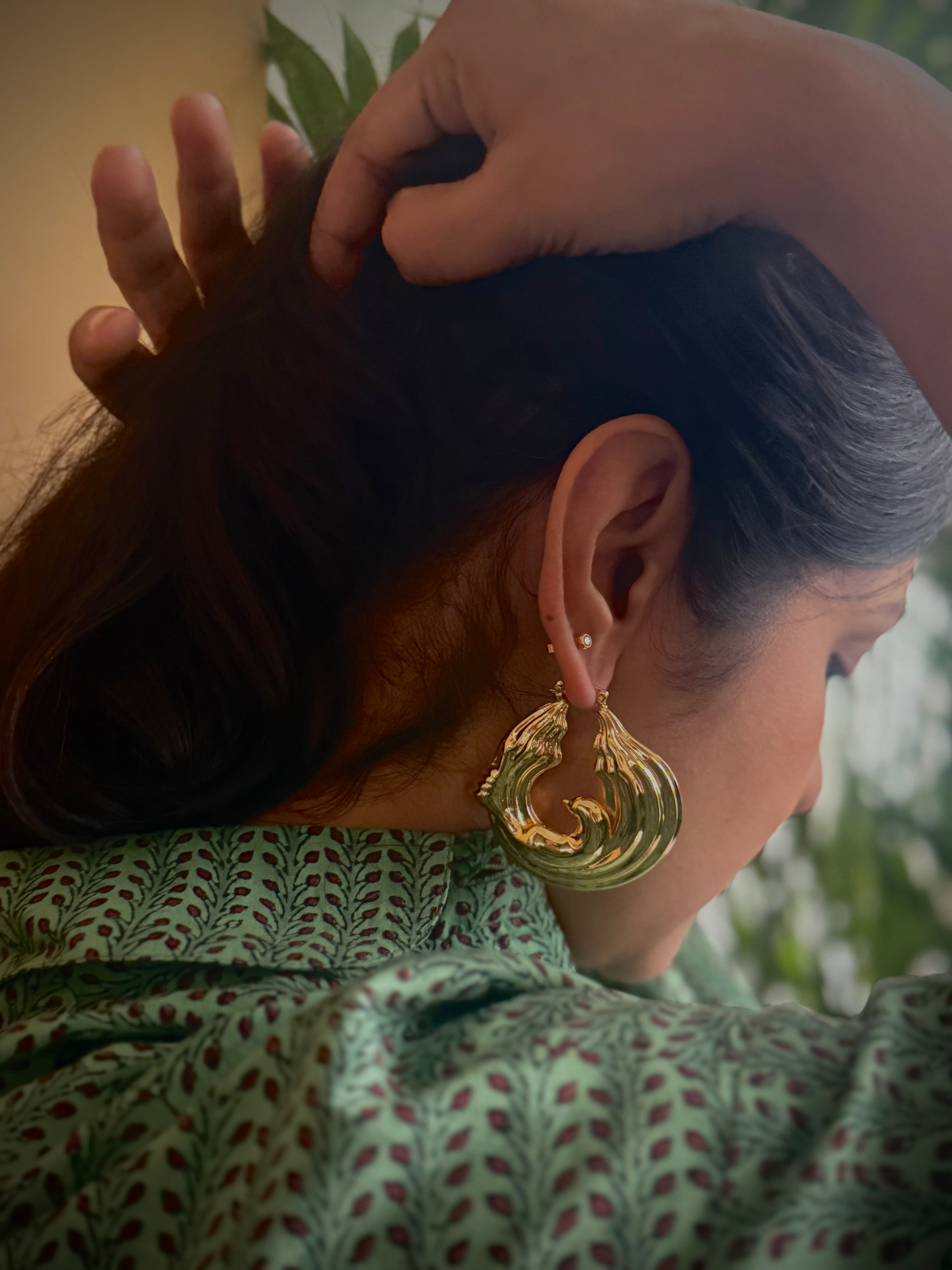Woman holding back her hair, wearing gold plated chunky hoop earrings with a green patterned top, surrounded by plants.