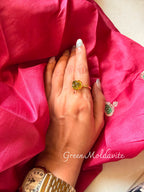 Green Moldavite ring with a faceted green stone held in gold prongs, worn on a hand resting on pink silk