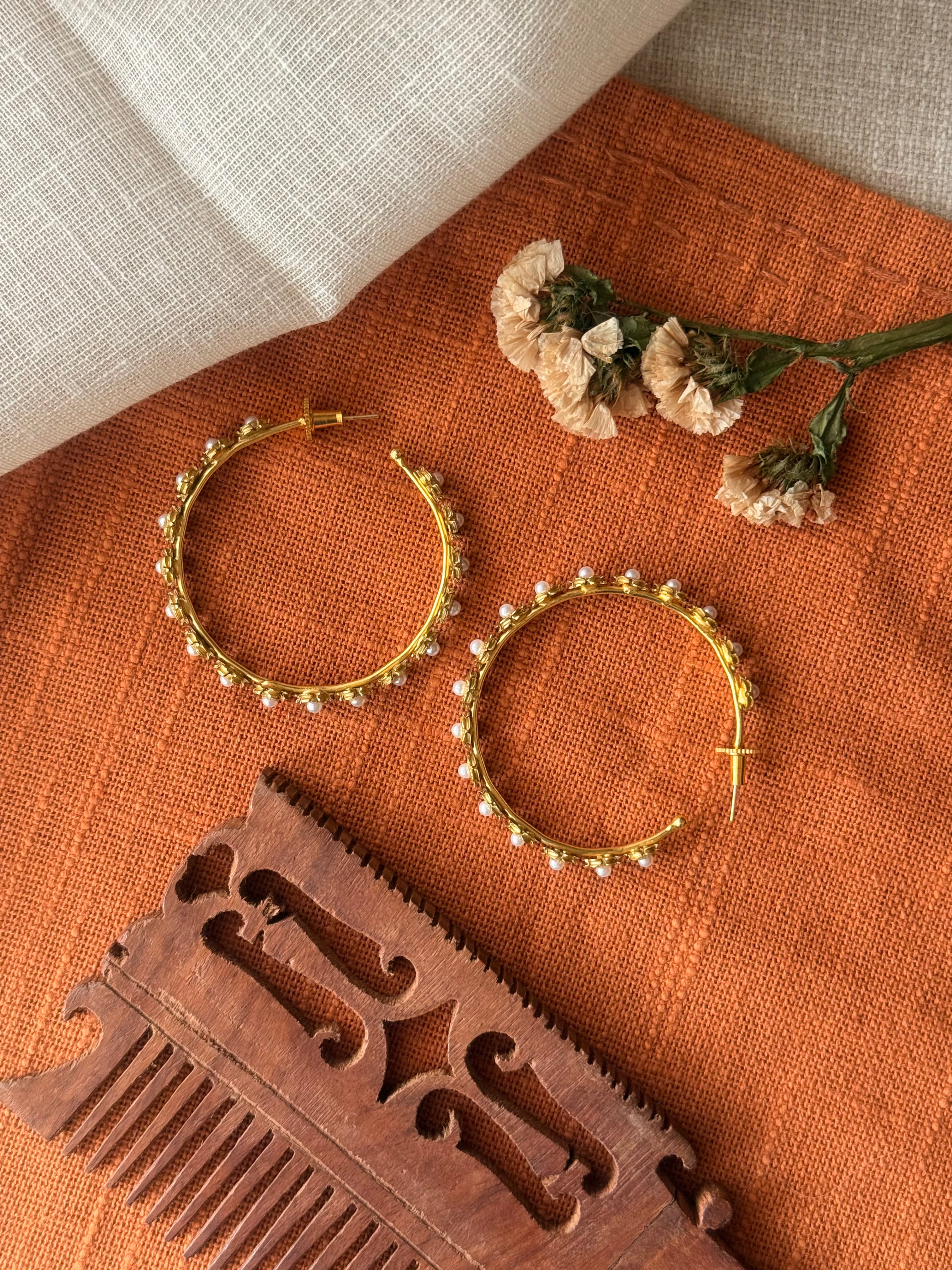 Flat lay of two Gulnaar Hoops on rust-orange linen, gold flower clusters with pearl centres along each hoop, styled with dried flowers and a carved wooden comb.