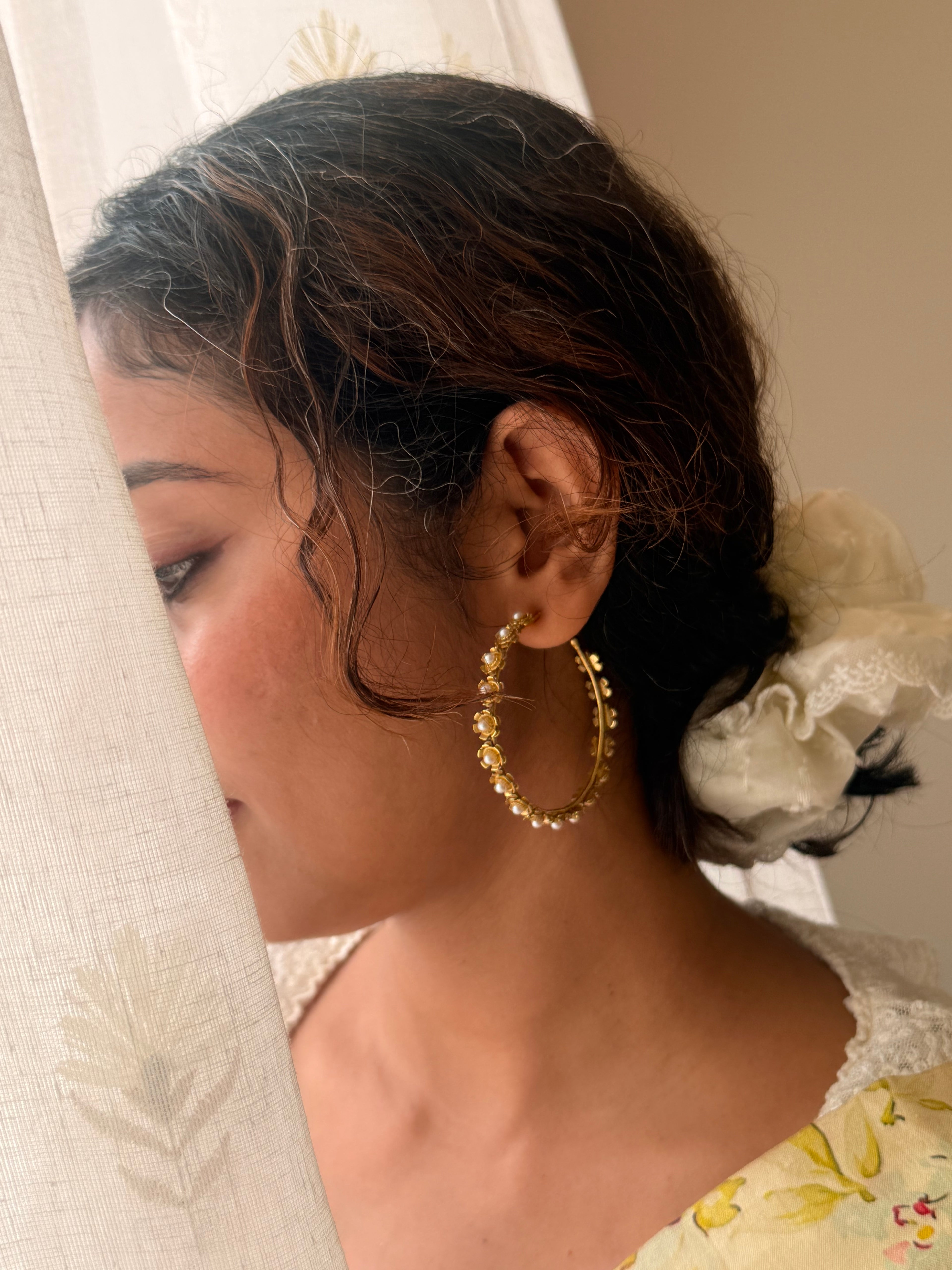 Gold-plated brass Gulnaar Hoops worn at the ear, small pearl-centred flower details along the hoop, hair swept back against an embroidered sheer curtain background.