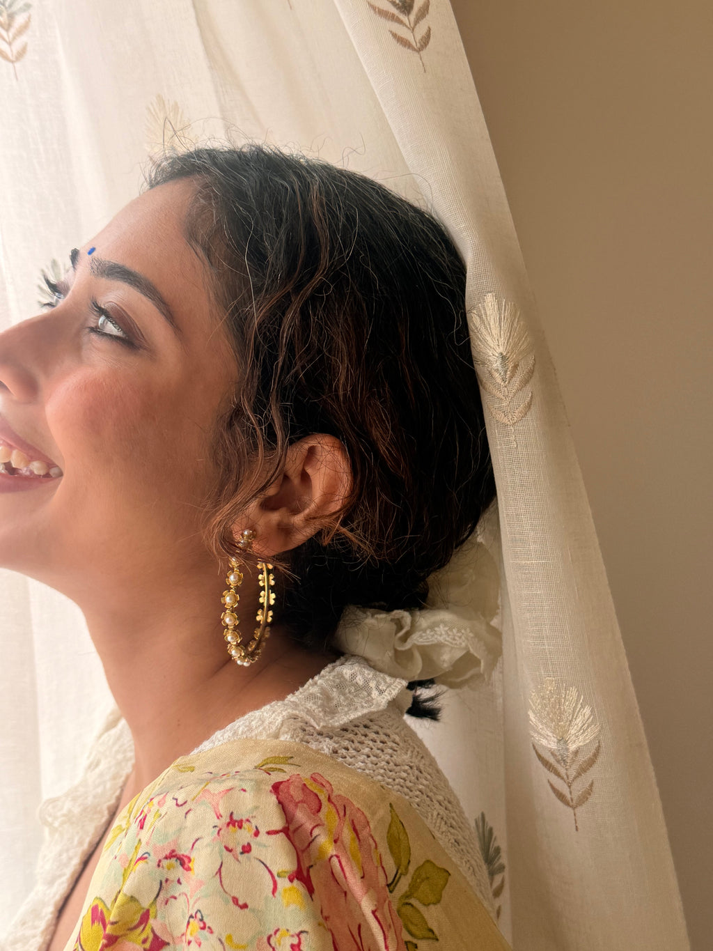 Woman smiling and looking upward, wearing Gulnaar Hoops with pearl-centred flower detail, sheer embroidered curtain in the background.